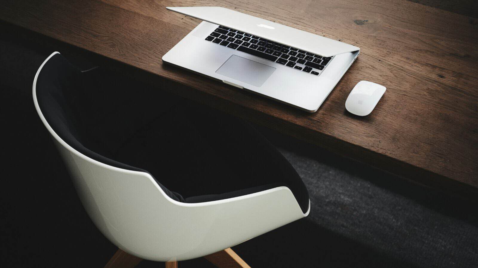 Laptop and wireless mouse on a wooden desk beside a white office chair