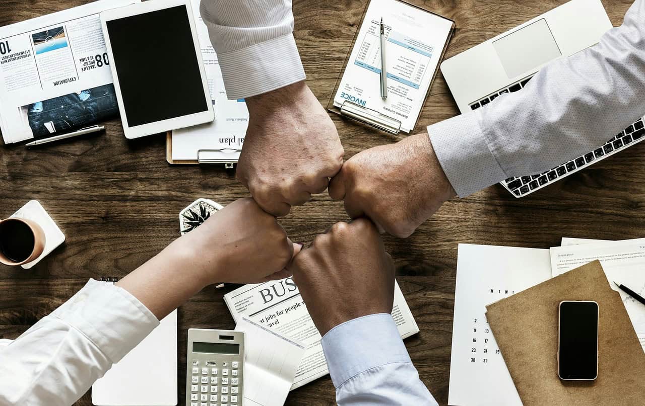 Team members fist bump over a cluttered desk with documents, tablets, and office supplies.